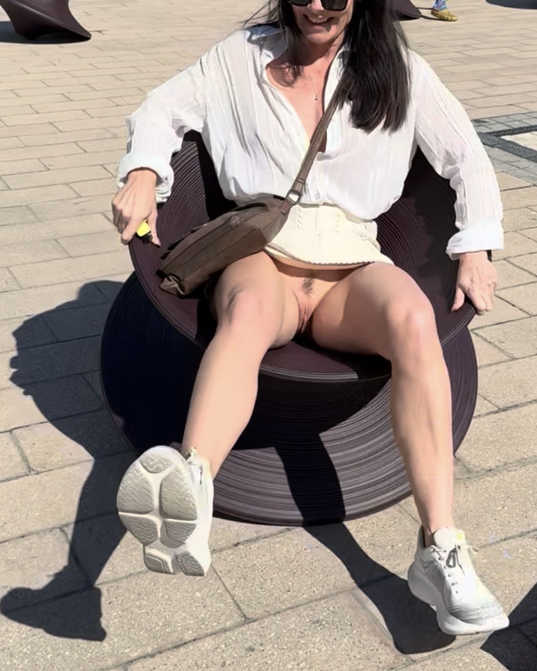 girl naked in public sneakers - Public Bench Exposure: Natural Bush on Display A woman sits on a public bench, partially nude with her natural bush exposed, wearing a mini skirt and white sneakers, smiling playfully. - Public Flashing Pics
