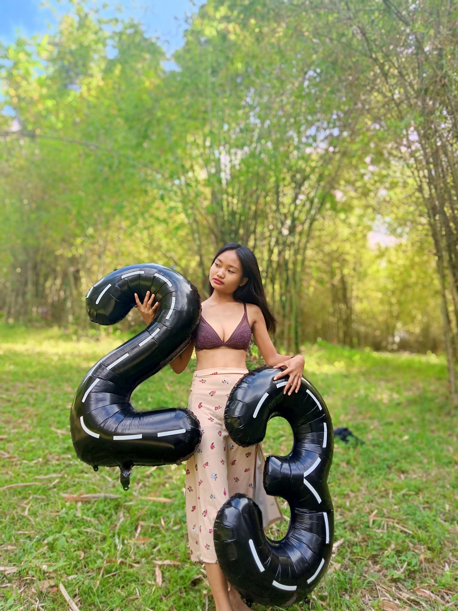 Hotwife Pics: What’s the Story Behind This Hotwife’s Public Display? A woman in a grassy area, wearing a dark purple lace bra and a light pink floral skirt, holds two large black number balloons, '2' and '3'.